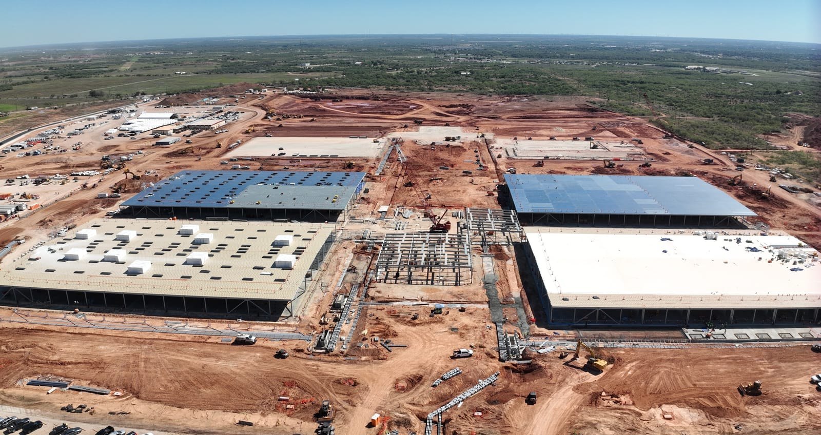 Actual Abilene data-center buildings during construction, with large roof sections, an unfinished central utility hall and extensive cleared red-soil ground.