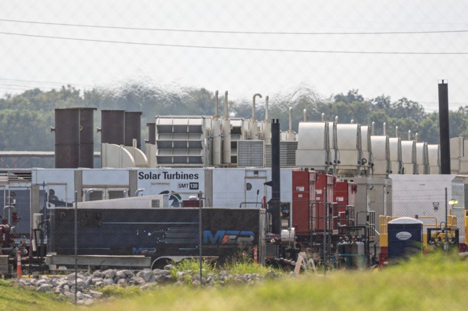 Roadside view of gas turbines at Colossus I behind chain-link fencing, with tall dark exhaust stacks.