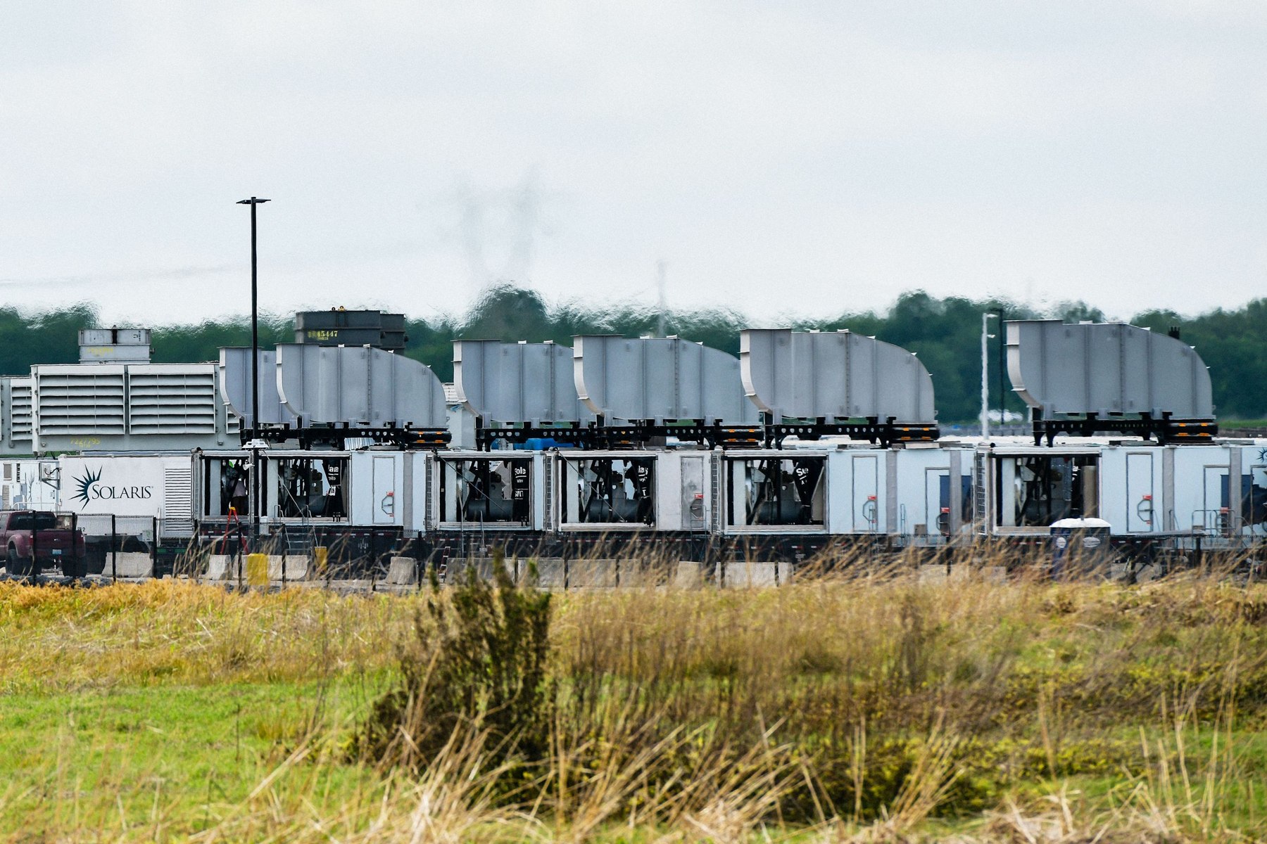 Ground-level gas turbine trailers at xAI’s Memphis facility behind a grassy verge, with visible exhaust stacks.